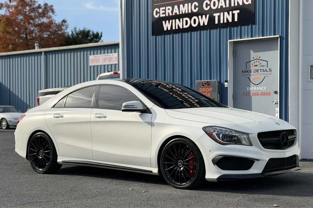 A white Mercedes-Benz CLA sedan with tinted windows, parked in front of a blue building with a sign for "Mike Details."